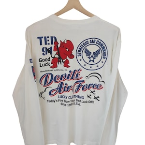 May include: Off-white long-sleeve shirt with a graphic design featuring a red devil, the text "Devil's Air Force," and the words "Strategic Air Command." The shirt is from Lucky Clothing.