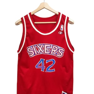 May include: A red basketball jersey with white and blue accents, hanging on a wooden hanger. The jersey features the word "SIXERS" in white and blue, and the number "42" in blue. A Champion logo is visible.