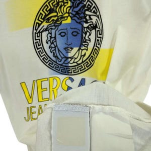 May include: Cream-colored t-shirt featuring a graphic of a Medusa head in a circular frame with yellow and blue accents. The word "VERSA" is printed in yellow, with "JEANS" below. The shirt's tag is visible.