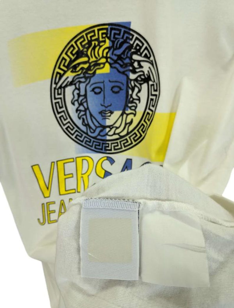 May include: Cream-colored t-shirt featuring a graphic of a Medusa head in a circular frame with yellow and blue accents. The word "VERSA" is printed in yellow, with "JEANS" below. The shirt's tag is visible.