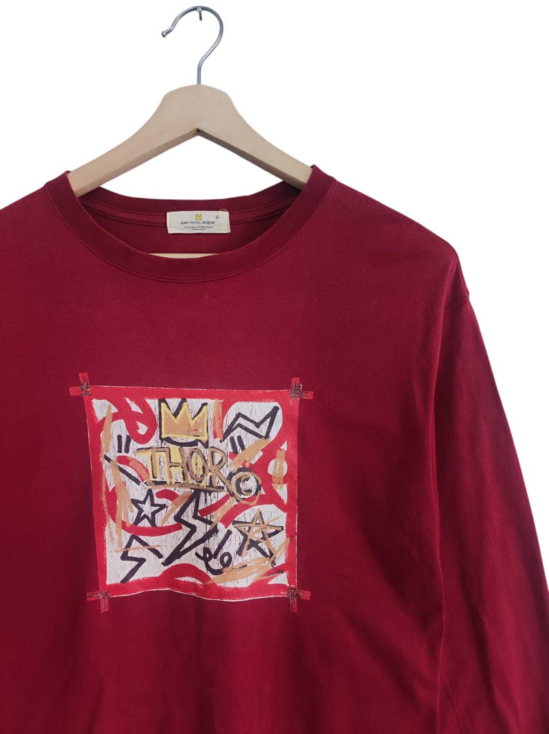 Puede incluir: Camiseta burdeos de manga larga con un dise&ntilde;o gr&aacute;fico. El dise&ntilde;o presenta una corona, estrellas y la palabra "THOR" en una est&eacute;tica de graffiti, enmarcada por un borde rojo. La camiseta est&aacute; colgada en una percha de madera.