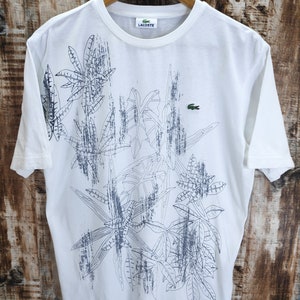 May include: White t-shirt with a crew neck and short sleeves. The shirt features a dark blue leaf design across the front and a small green crocodile logo. The brand name "Lacoste" is visible on the label.