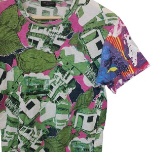 Może przedstawiać: T-shirt z kr&oacute;tkim rękawem o efektownym, abstrakcyjnym wzorze. Wz&oacute;r zawiera zielone, białe i czarne kształty, przypominające liście i budynki, na r&oacute;żowym tle. Rękaw ma kolorowy, artystyczny design.