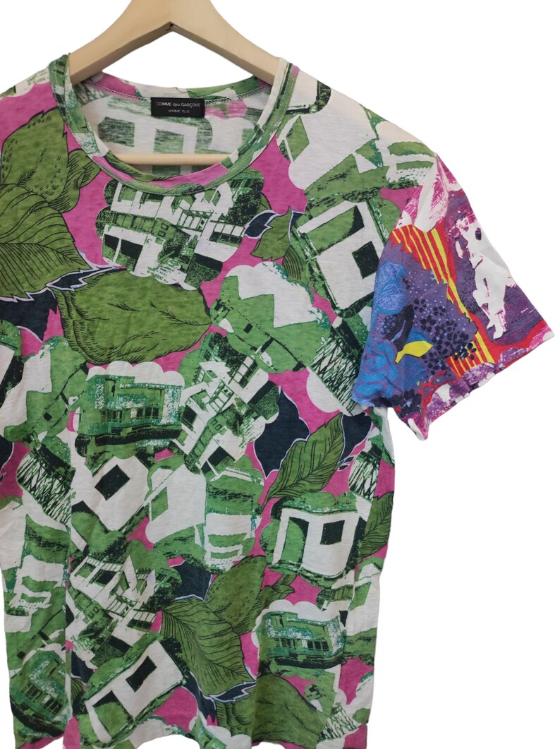 Może przedstawiać: T-shirt z kr&oacute;tkim rękawem o efektownym, abstrakcyjnym wzorze. Wz&oacute;r zawiera zielone, białe i czarne kształty, przypominające liście i budynki, na r&oacute;żowym tle. Rękaw ma kolorowy, artystyczny design.