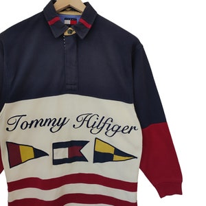 May include: A long-sleeved Tommy Hilfiger rugby shirt. The shirt features a navy blue collar and sleeves, a white midsection with the brand name and nautical flags, and red accents. The bottom half has red and white stripes.