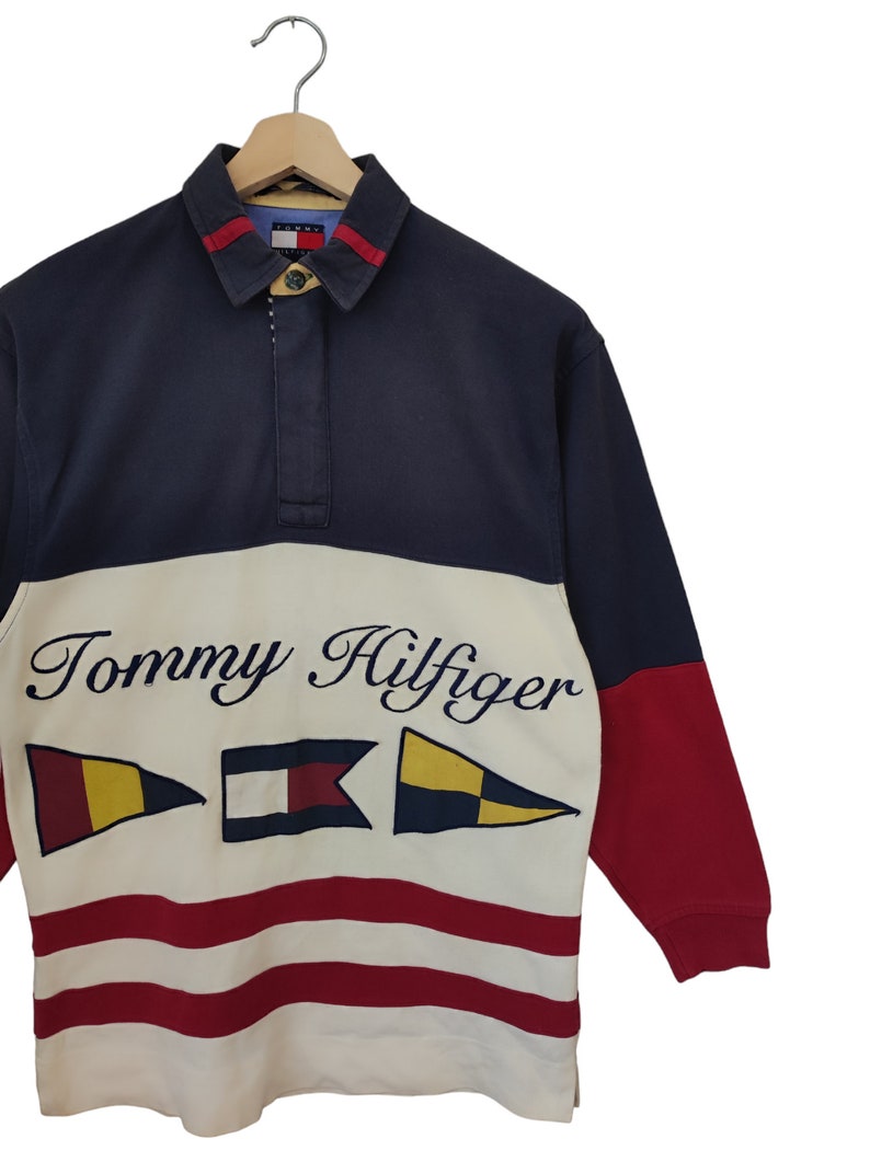May include: A long-sleeved Tommy Hilfiger rugby shirt. The shirt features a navy blue collar and sleeves, a white midsection with the brand name and nautical flags, and red accents. The bottom half has red and white stripes.