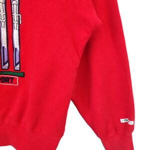 May include: A red sweatshirt with a ski-themed design. The design features two pairs of skis in white, purple, and green, with the word "SPORT" in a white box. The sleeve has a small white tag with text.