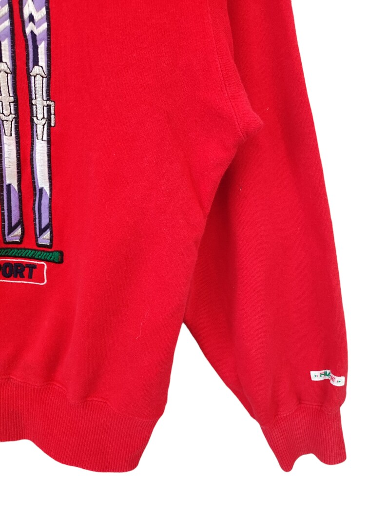 May include: A red sweatshirt with a ski-themed design. The design features two pairs of skis in white, purple, and green, with the word "SPORT" in a white box. The sleeve has a small white tag with text.