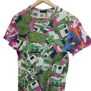 Może przedstawiać: T-shirt z kr&oacute;tkim rękawem o żywym, abstrakcyjnym wzorze. Koszulka ma r&oacute;żowe tło z zielonymi liśćmi, białymi geometrycznymi kształtami i kolorowymi akcentami na rękawach. Koszulka wisi na drewnianym wieszaku.