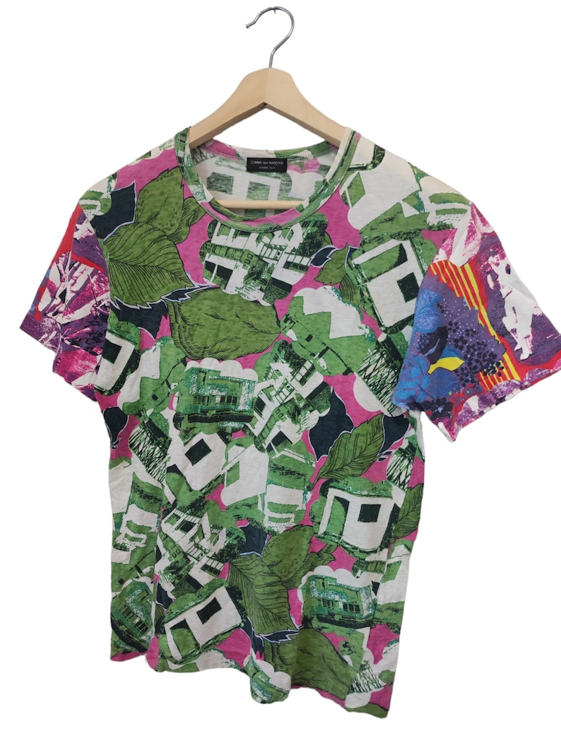 Może przedstawiać: T-shirt z kr&oacute;tkim rękawem o żywym, abstrakcyjnym wzorze. Koszulka ma r&oacute;żowe tło z zielonymi liśćmi, białymi geometrycznymi kształtami i kolorowymi akcentami na rękawach. Koszulka wisi na drewnianym wieszaku.