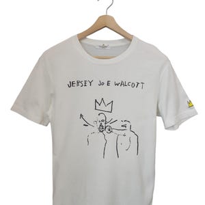 Puede incluir: Camiseta blanca con el texto "JERSEY JOE WALCOTT" sobre una ilustraci&oacute;n en blanco y negro de dos figuras con una corona. La manga derecha tiene un dise&ntilde;o amarillo y negro. La camiseta est&aacute; colgada en una percha de madera.