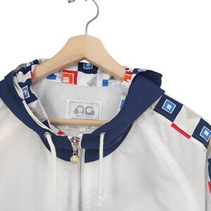 May include: White hooded jacket with a navy blue trim and geometric pattern. The jacket features a full zipper, drawstring, and a logo on the inside. The jacket is hanging on a wooden hanger.