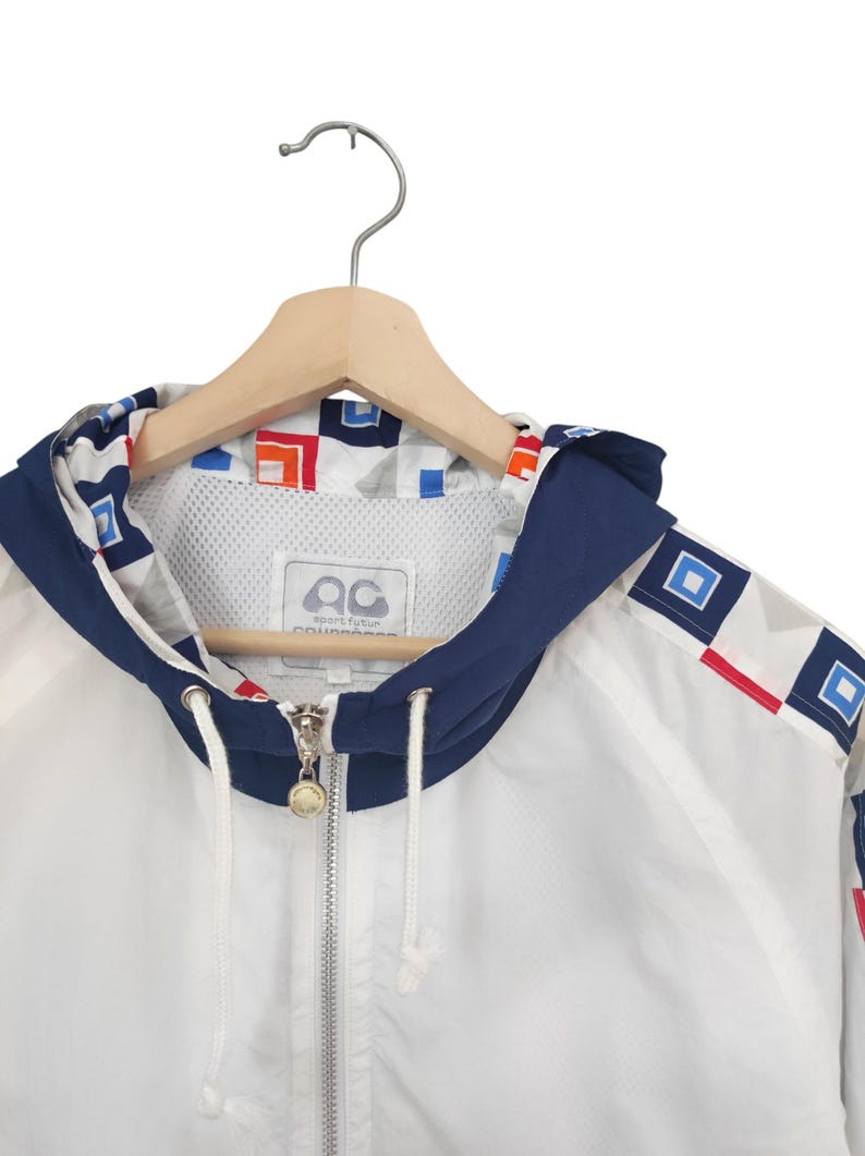 May include: White hooded jacket with a navy blue trim and geometric pattern. The jacket features a full zipper, drawstring, and a logo on the inside. The jacket is hanging on a wooden hanger.