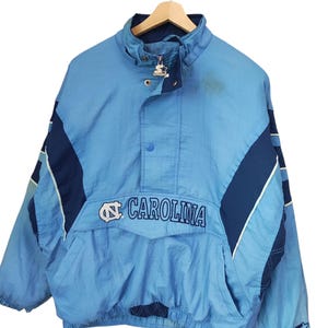 May include: A light blue and navy blue pullover windbreaker jacket featuring the text CAROLINA and the North Carolina Tar Heels logo on the front. The jacket has a high collar with a zip closure, a snap button placket, and a Starter logo.