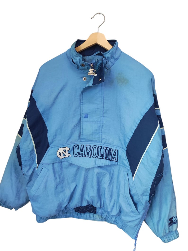 May include: A light blue and navy blue pullover windbreaker jacket featuring the text CAROLINA and the North Carolina Tar Heels logo on the front. The jacket has a high collar with a zip closure, a snap button placket, and a Starter logo.