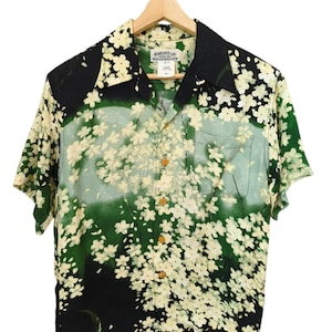 Puede incluir: Camisa de manga corta con botones y estampado floral. La camisa tiene cuello negro y un bolsillo. El diseño presenta flores blancas sobre un fondo verde y negro. La camisa está colgada de una percha de madera.