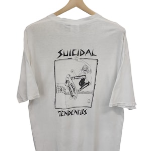 May include: White t-shirt with a black graphic of a skateboarder and the words "SUICIDAL TENDENCIES". The shirt is hanging on a wooden hanger.