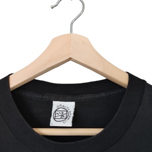 May include: Black crew neck t-shirt on a wooden hanger. The shirt features a white tag with the text "GRAVITY GRAPHICS" and a logo. The shirt is size L.