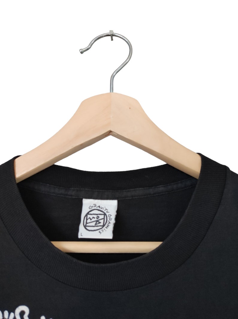 May include: Black crew neck t-shirt on a wooden hanger. The shirt features a white tag with the text "GRAVITY GRAPHICS" and a logo. The shirt is size L.