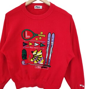 May include: Red FILA Sport sweatshirt with embroidered sports equipment graphics. Features a tennis racket, skis, golf club, and ski boots. The text "BIELLA ITALIA" is also embroidered on the front.