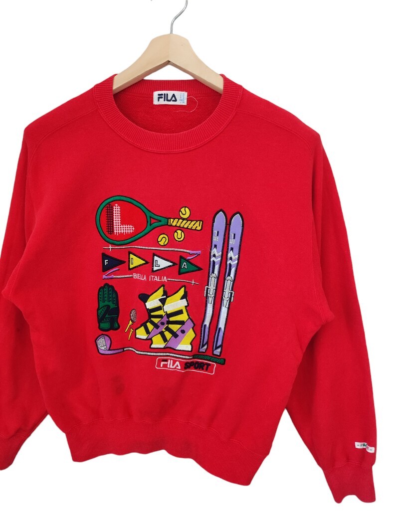 May include: Red FILA Sport sweatshirt with embroidered sports equipment graphics. Features a tennis racket, skis, golf club, and ski boots. The text "BIELLA ITALIA" is also embroidered on the front.