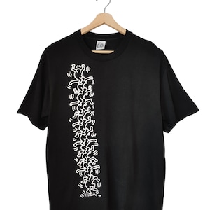 May include: Black t-shirt with a white graphic of Keith Haring figures in a vertical line. The figures are simple, stick-figure-like people. The t-shirt is displayed on a wooden hanger.