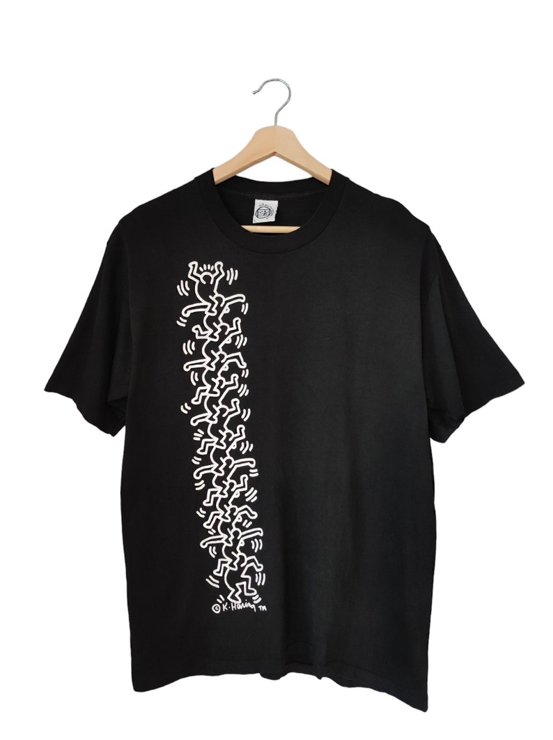 May include: Black t-shirt with a white graphic of Keith Haring figures in a vertical line. The figures are simple, stick-figure-like people. The t-shirt is displayed on a wooden hanger.