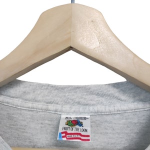 May include: A light gray Fruit of the Loom t-shirt, size XL, hanging on a wooden hanger. The shirt's tag, featuring the Fruit of the Loom logo and "Made in U.S.A.", is visible. The shirt is made of a soft, likely cotton blend material.