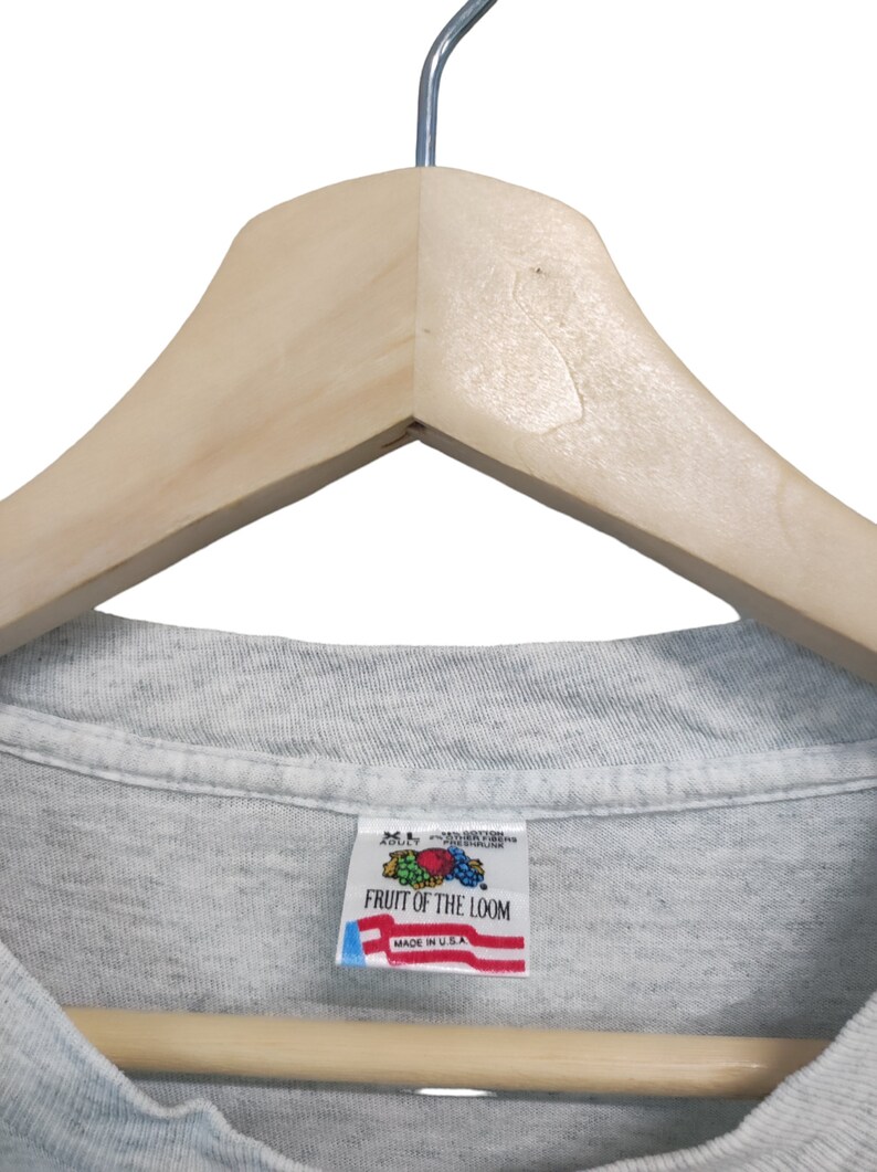 May include: A light gray Fruit of the Loom t-shirt, size XL, hanging on a wooden hanger. The shirt's tag, featuring the Fruit of the Loom logo and "Made in U.S.A.", is visible. The shirt is made of a soft, likely cotton blend material.