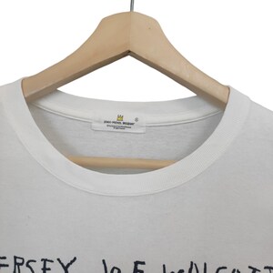 Puede incluir: Camiseta blanca de cuello redondo con texto negro "JERSEY JOE WALCOTT". La camiseta est&aacute; colgada de una percha de madera. La etiqueta dice "JEAN-MICHEL BASQUIAT".