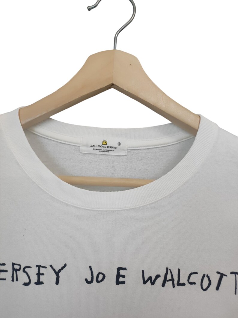 Puede incluir: Camiseta blanca de cuello redondo con texto negro "JERSEY JOE WALCOTT". La camiseta est&aacute; colgada de una percha de madera. La etiqueta dice "JEAN-MICHEL BASQUIAT".