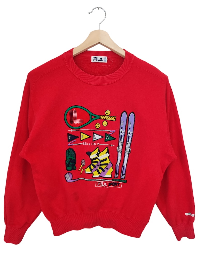 May include: Red Fila sweatshirt with a graphic of sports equipment including a tennis racket, skis, and golf club. The sweatshirt has the Fila logo on the front and the sleeve.