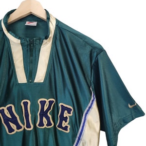 May include: Teal and cream-colored Nike athletic top with a quarter-zip closure. The word "NIKE" is embroidered in navy blue with a cream outline. A small Nike swoosh logo is on the sleeve. The top is hanging on a wooden hanger.