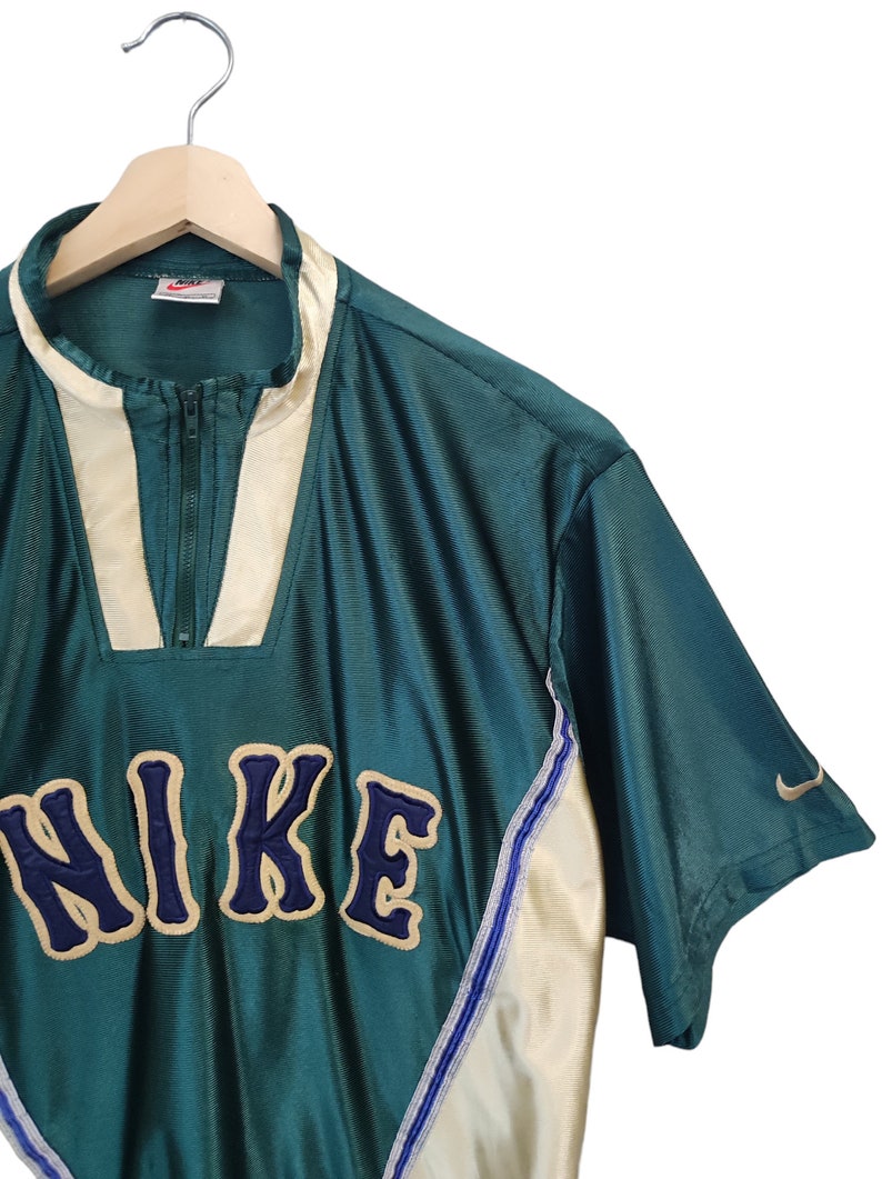 May include: Teal and cream-colored Nike athletic top with a quarter-zip closure. The word "NIKE" is embroidered in navy blue with a cream outline. A small Nike swoosh logo is on the sleeve. The top is hanging on a wooden hanger.