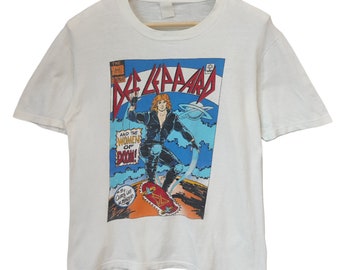 Puede incluir: Camiseta blanca con un gráfico estilo cómic. El gráfico incluye el nombre de la banda "Def Leppard" en rojo y azul, y una ilustración de una persona en monopatín. El texto en el gráfico dice "And the Women of Doom!"