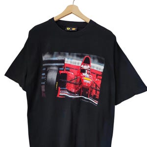 May include: Black t-shirt featuring a close-up image of a red Formula 1 race car in motion. The car has a prominent black tire and is set against a blurred background. The text "POWERING THE DREAM" is printed at the bottom.