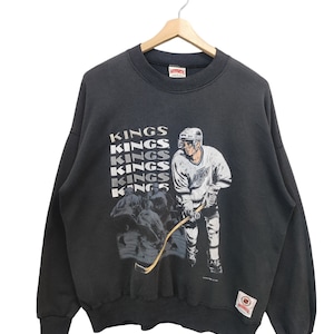 K&ouml;nnte beinhalten: Schwarzes Vintage Kings Hockey-Sweatshirt mit einer Grafik eines Hockeyspielers und dem Wort "KINGS" vertikal wiederholt. Das Sweatshirt hat einen Rundhalsausschnitt und lange &Auml;rmel. Ein Aufn&auml;her ist unten rechts sichtbar.