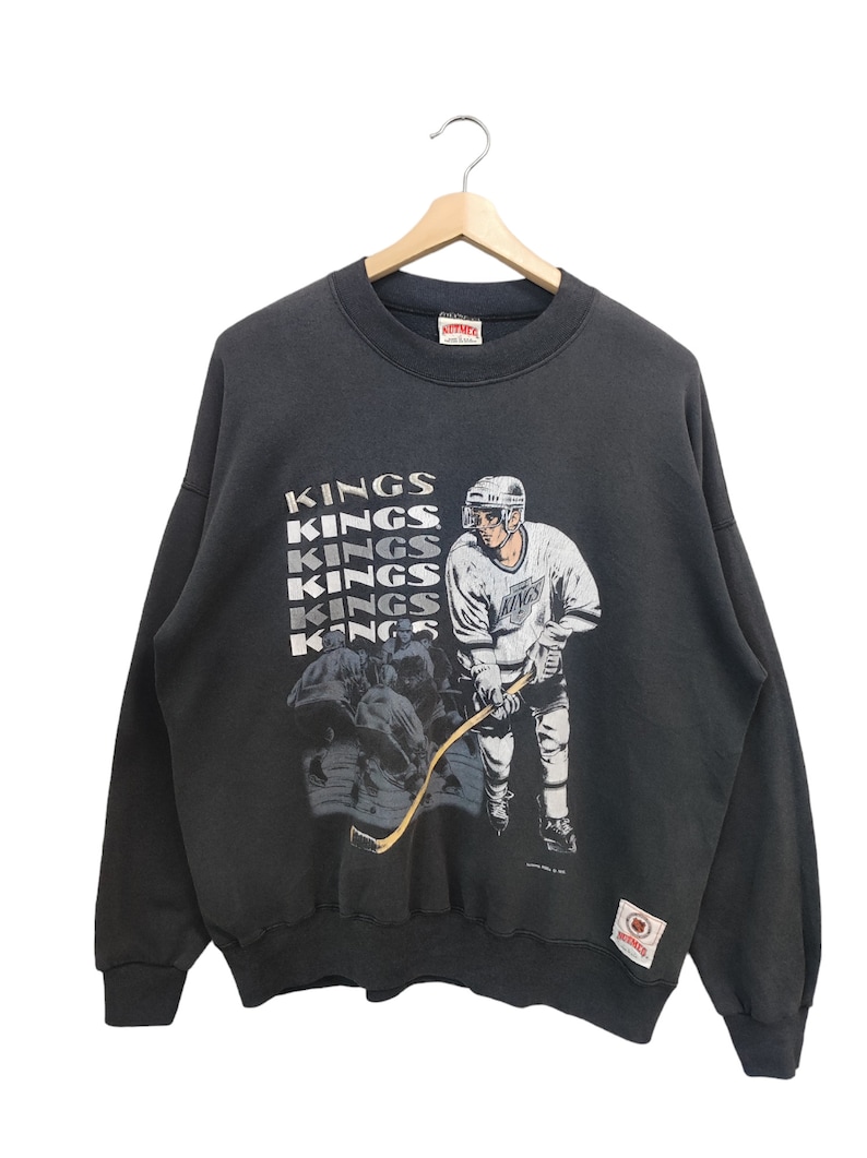 K&ouml;nnte beinhalten: Schwarzes Vintage Kings Hockey-Sweatshirt mit einer Grafik eines Hockeyspielers und dem Wort "KINGS" vertikal wiederholt. Das Sweatshirt hat einen Rundhalsausschnitt und lange &Auml;rmel. Ein Aufn&auml;her ist unten rechts sichtbar.