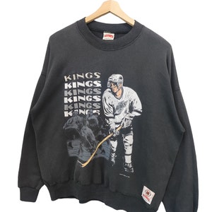 K&ouml;nnte beinhalten: Ein schwarzes Vintage-Sweatshirt mit einer Hockey-Spieler-Grafik und dem Wort "KINGS" vertikal wiederholt. Die Grafik zeigt einen Hockeyspieler in einem wei&szlig;en Trikot und ein Teamlogo. Das Sweatshirt h&auml;ngt an einem Holzkleiderb&uuml;gel.