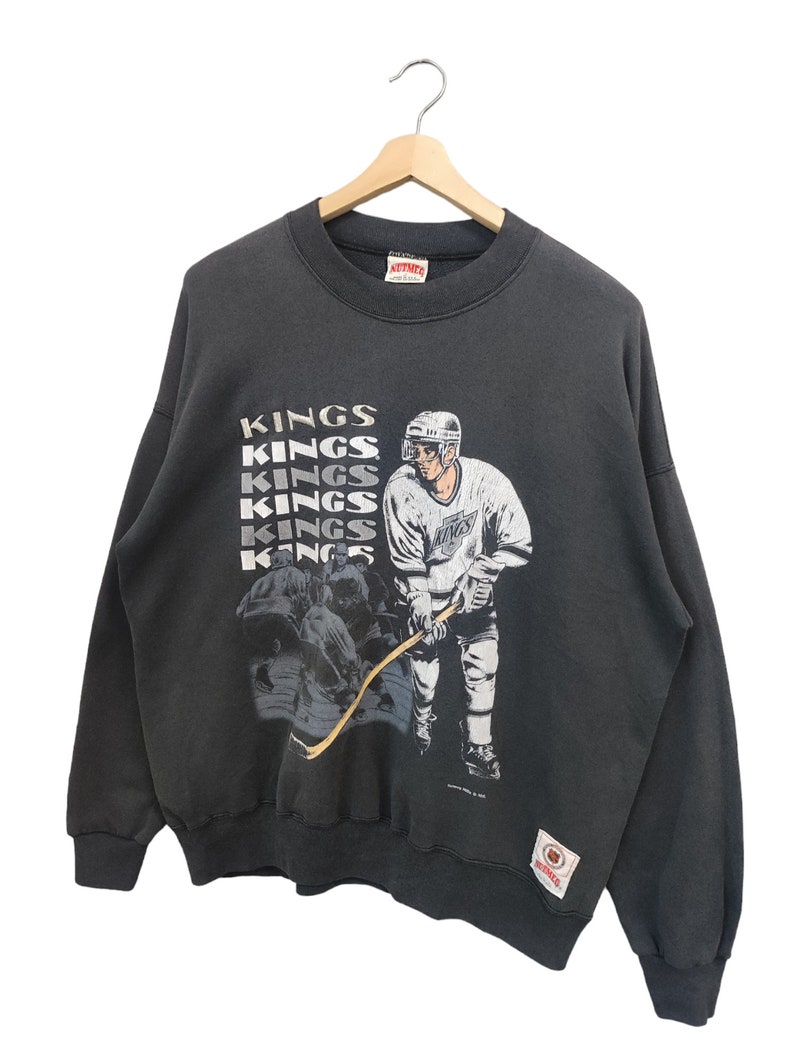 K&ouml;nnte beinhalten: Ein schwarzes Vintage-Sweatshirt mit einer Hockey-Spieler-Grafik und dem Wort "KINGS" vertikal wiederholt. Die Grafik zeigt einen Hockeyspieler in einem wei&szlig;en Trikot und ein Teamlogo. Das Sweatshirt h&auml;ngt an einem Holzkleiderb&uuml;gel.