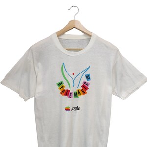 以下が含まれることがあります： カラフルな「Apple Magic」グラフィックがプリントされた白いTシャツ。デザインには、様式化された人物、Appleのロゴ、そして年号「95」が含まれています。Tシャツは木製のハンガーに掛けられています。