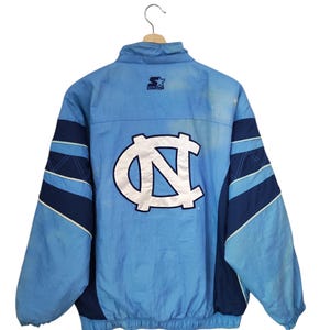 May include: A light blue and navy blue Starter brand windbreaker jacket featuring a large white North Carolina Tar Heels logo on the back. The jacket has a high collar, long sleeves with navy blue stripes, and an elasticated hem with a drawstring.