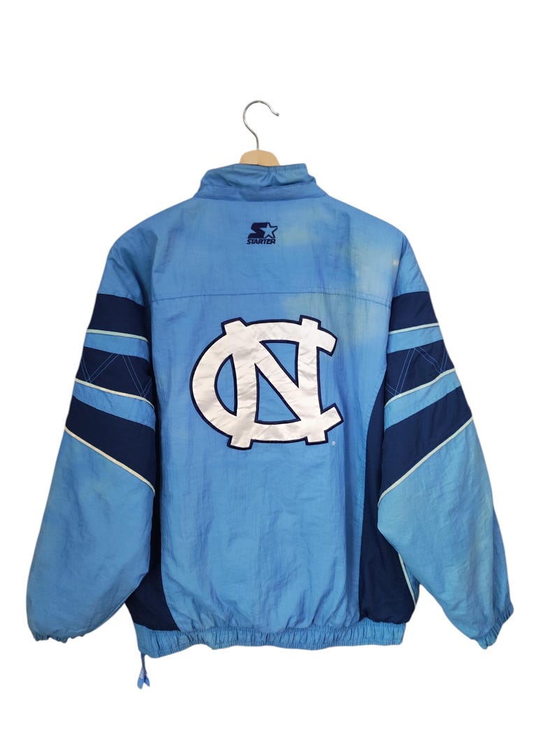 May include: A light blue and navy blue Starter brand windbreaker jacket featuring a large white North Carolina Tar Heels logo on the back. The jacket has a high collar, long sleeves with navy blue stripes, and an elasticated hem with a drawstring.