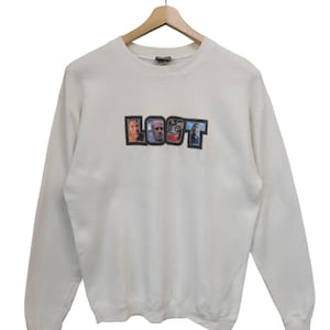 May include: Cream-colored sweatshirt with the word "LOST" in a rectangular design. The word "LOST" features images of people. The sweatshirt is hanging on a wooden hanger.