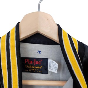 May include: Black and yellow varsity jacket with a black collar and yellow stripes. The jacket is hanging on a wooden hanger. The label reads "Pla-Jac by Dunbrooke" and "Made in USA".