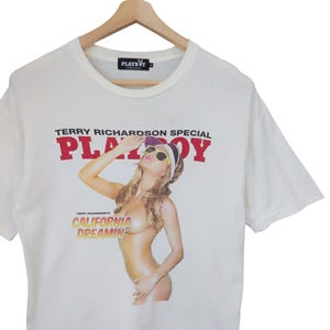 May include: White t-shirt featuring a graphic of a woman wearing sunglasses and a visor, with the text "TERRY RICHARDSON SPECIAL PLAYBOY" in red. Below the image is the text "CALIFORNIA DREAMIN'". The shirt is hanging on a wooden hanger.