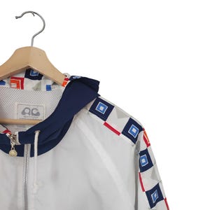 May include: White zip-up jacket with a navy blue hood and geometric pattern accents in red, blue, and orange. The jacket is hanging on a wooden hanger. The jacket has a logo on the chest.