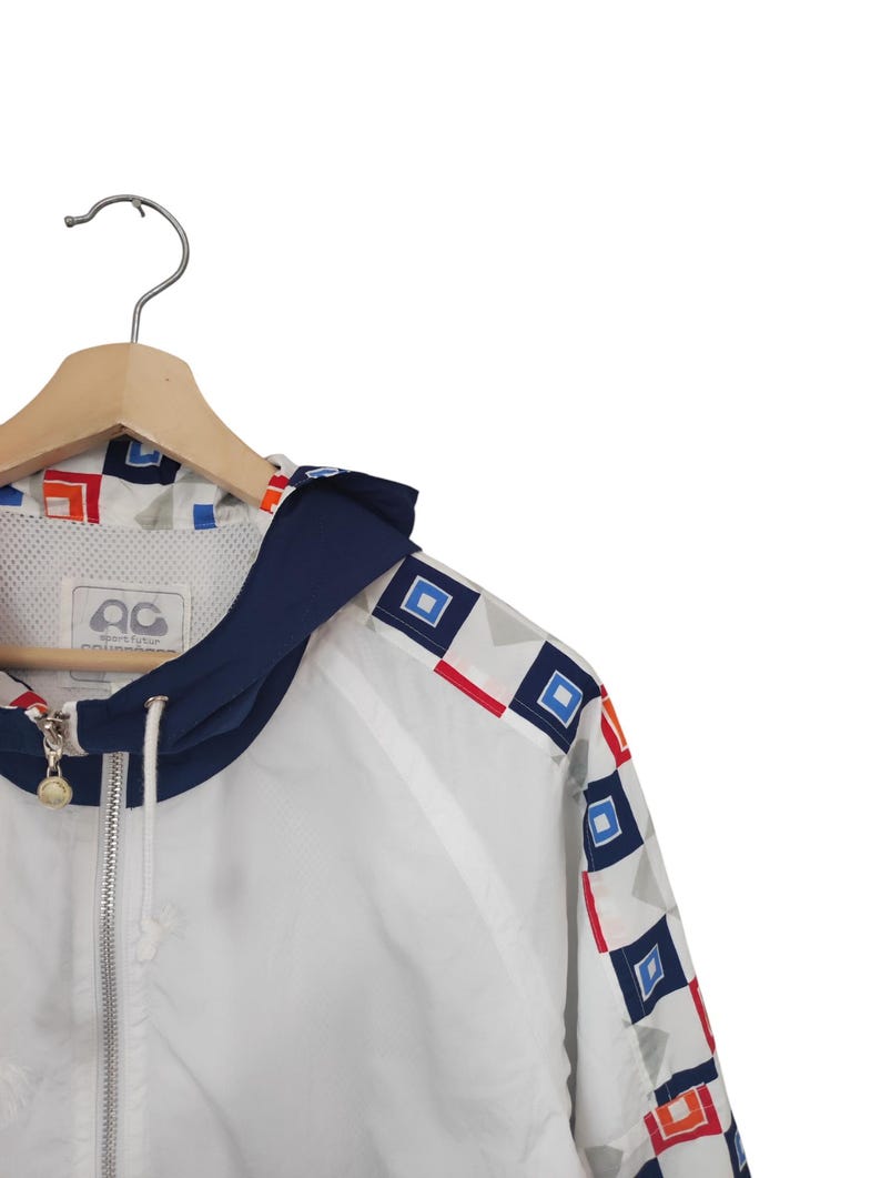 May include: White zip-up jacket with a navy blue hood and geometric pattern accents in red, blue, and orange. The jacket is hanging on a wooden hanger. The jacket has a logo on the chest.