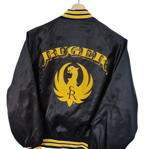May include: Black satin jacket with yellow and black striped trim. The back features the word "RUGER" in yellow arched lettering above a yellow phoenix logo. The jacket is hanging on a wooden hanger.