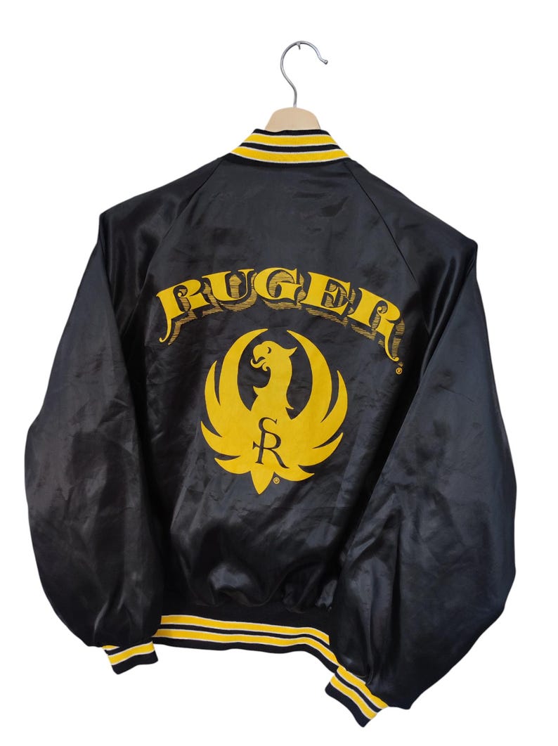 May include: Black satin jacket with yellow and black striped trim. The back features the word "RUGER" in yellow arched lettering above a yellow phoenix logo. The jacket is hanging on a wooden hanger.